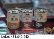 Original souvenirs from St. Petersburg — tinned jars with the inscription “Land of St. Petersburg,” decorated in the style of gift wrapping. In the foreground are three jars with different names associated with famous places in the city: “Petropavlovka Beach,” “V.O. Spit,” and “Summer Garden.” Each jar has its own color scheme and illustration: the silhouette of the Peter and Paul Fortress, a building on the Spit of Vasilievsky Island, and the alleys of the Summer Garden. These souvenirs symbolically contain soil from the indicated landmarks and serve as an unusual memorable gift for tourists. In the background, other souvenirs are visible — badges, magnets, and keychains. (2014 год). Редакционное фото, фотограф Анна Кривицкая / Фотобанк Лори