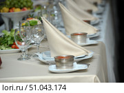 An elegantly laid festive table. In the foreground are neatly folded cream-colored fabric napkins in the shape of a pyramid, placed on white plates. In front of each napkin is a small metal bowl filled with red caviar, placed on a separate saucer with a fork and napkin. Nearby are empty, overturned wine or champagne glasses. In the depths of the table, you can see plates with greens, vegetables, fruits, as well as other served places, repeating the composition. The light-colored tablecloth creates an atmosphere of solemnity, and the soft lighting gives the frame warmth and a festive character. Стоковое фото, фотограф Анна Кривицкая / Фотобанк Лори