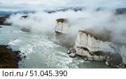 River with white water rapids flowing alongside high white cliffs obscured by dense fog and clouds below mountains. Стоковое AI-изображение, AI-художник Анжелика Самсонова / Фотобанк Лори