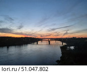A picturesque view of the river in the evening. On the horizon are the warm shades of the sunset, smoothly transitioning from bright orange and pink to a deep blue sky. Light cirrus clouds stretch across the sky, creating the effect of soft brushstrokes. In the center of the frame you can see the bridge connecting the banks, its dark silhouette clearly stands out against the glowing horizon. On the banks of the river and on the bridge itself, lights are lit, reflecting in the calm water surface. In the right part of the foreground is the dark silhouette of a cliff with vegetation. The atmosphere of the picture conveys the peace and beauty of the sunset hour. Стоковое фото, фотограф Анна Кривицкая / Фотобанк Лори