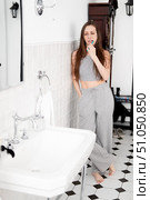 A beautiful and young woman in gray pajamas with braces stands in the bathroom leaning against the wall and brushing her teeth. Стоковое фото, фотограф Кашапова Эльвира / Фотобанк Лори