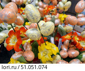A composition of decorative artificial flowers and eggs. Flowers of bright shades - yellow and orange-red - are made of synthetic materials, with detailed petals and cores. Between them are decorative eggs, painted in delicate pastel tones - yellow and peach - with a silver spray effect, imitating an aged or metallic surface. The stems of the decorations are green, with artificial leaves. The composition conveys the atmosphere of a spring holiday and Easter theme, creating a bright and festive mood. Стоковое фото, фотограф Анна Кривицкая / Фотобанк Лори