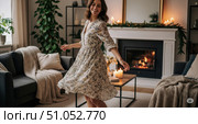 Cozy home lifestyle with happy woman dancing in living room, beautiful smiling female in floral dress enjoying comfortable evening by fireplace with candles. Стоковое AI-изображение, AI-художник Павел Муравьев / Фотобанк Лори