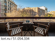 A cozy balcony scene with two wooden chairs and a small table. Two blue cups sit on the table. Urban buildings and trees are visible in the background. Стоковое фото, фотограф PAv / Фотобанк Лори