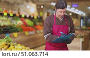 Man store employee with documents takes inventory of goods on display case of vegetable store. Seller records quantity of goods in report, draws up document on availability of vegetables in store. Стоковое видео, видеограф Яков Филимонов / Фотобанк Лори