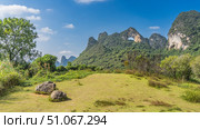 Beautiful mountain landscape. A cliff with steep slopes and a rounded arched hole at the top. Green vegetation, boulders in the meadow. Blue sky, clouds.A sunny day. China. Yangshuo. Moon Hill. Стоковое фото, фотограф Вера Ивановна Кубарева / Фотобанк Лори
