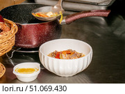 Белая керамическая миска с горячим супом, наполненным овощами и морепродуктами, стоит на металлической кухонной поверхности. Рядом небольшая белая пиала с соусом или приправой. На заднем плане повар черпаком наливает суп из красной кастрюли с покрытием. Слева частично видна плетёная корзинка с продуктами. Стоковое фото, фотограф Ксения Чунаева / Фотобанк Лори