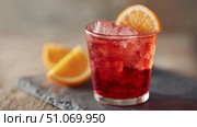 This vibrant cocktail presents a captivating blend of deep ruby red tones and glistening ice, creating a visually striking drink that promises a refreshing experience. The chilled glass enhances the coolness, with condensation hinting at the crispness of the first sip. The drink is elegantly garnished with a fresh, juicy slice of orange, adding both a pop of warm color and a hint of citrus aroma that complements the rich hue of the beverage. In the background, wedges of orange rest on a natural slate surface, subtly suggesting the fresh ingredients and artisans preparation behind the drink. The overall presentation strikes a perfect balance between sophistication and casual enjoyment, making it ideal for warm-weather gatherings, upscale events, or simply savoring a moment of indulgence. Стоковое AI-изображение, AI-художник Ксения Чунаева / Фотобанк Лори