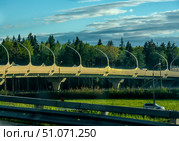 A modern automobile bridge in St. Petersburg with unique curved noise barrier structures, rising on strong supports. The bridge passes over a green area with a dense forest in the background, creating a contrast between the engineering architecture and the natural landscape. In the foreground, elements of the road barriers and a car moving along the lower level of the road are visible. The warm light of the sunset or morning sun softly illuminates the bridge and gives a golden hue to its structure. In the sky, there is a combination of blue tones and clouds, creating a picturesque background. Стоковое фото, фотограф Анна Кривицкая / Фотобанк Лори
