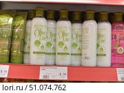 A shelf in a hair care store with bottles and tubes of Olive Line and Dalan d’Olive conditioner lined up. In the center are white plastic bottles of Olive Line hair conditioner, 400 ml, with green caps and a minimalist label design featuring olives and a drop of oil. The label states that the product is suitable for all hair types and contains olive oil. On the left are tubes of Dalan d’Olive conditioner in green packaging. In the foreground is a price tag with information about the product and its cost. The photo was taken in a store with bright lighting, with the focus on the central bottles. (2015 год). Редакционное фото, фотограф Анна Кривицкая / Фотобанк Лори