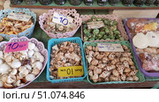 A souvenir counter with various keychains made of natural shells laid out in decorative plastic baskets. Each basket contains shells of different sizes, shapes and colors: white, cream, brown, motley, green. The keychains are equipped with metal rings for keys. The baskets have price tags indicating the cost in Thai baht - 10 or 20 baht. The background consists of a wooden table surface and a shelf with stone souvenirs. The composition conveys the atmosphere of a tourist market in coastal regions. Стоковое фото, фотограф Анна Кривицкая / Фотобанк Лори