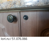 A close-up of wooden kitchen cabinets under a stone countertop. On the left, the door has a bronze-colored metal handle with a decorative pattern along the edge. On the right, there is a door without a handle, in place of which you can see a protruding screw with a washer, ready for installation or replacement of fittings. The surface of the cabinets is made to look like light wood with a pronounced texture, and the countertop has a marble pattern in beige and brown tones. The image conveys the moment of repair or replacement of furniture handles. Стоковое фото, фотограф Анна Кривицкая / Фотобанк Лори