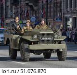 A military parade in the city. In the foreground is a historic army vehicle from the Second World War, painted olive green. On the front bumper is a sign with the inscription LENRESERVE. Behind the wheel is a man in modern clothes, next to him and in the back seat are two women in the uniform of the Great Patriotic War, one of whom is in a leather jacket, the other in a tunic with a cap. They smile and greet the spectators. In the background is a city street with historical architecture, flags and spectators along the road. (2015 год). Редакционное фото, фотограф Анна Кривицкая / Фотобанк Лори