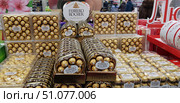 A supermarket display case with a large assortment of Ferrero Rocher chocolates, neatly laid out in transparent plastic packages of various formats. The candies are wrapped in gold foil, creating an effect of luxury and a festive mood. In the center of the composition are round and rectangular boxes with the Ferrero Rocher brand logo, and in the background is a decorative stand with a large brand name. The background includes elements of the sales area with customers and other goods, creating an atmosphere of pre-New Year or holiday shopping. (2019 год). Редакционное фото, фотограф Анна Кривицкая / Фотобанк Лори