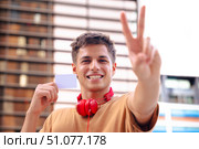 Smiling young man with headphones showing blank card and making peace sign outdoors. Стоковое фото, фотограф Raquel Corcoles Mendez / Фотобанк Лори