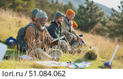 A senior African man with a beard sits on grass, reading a map. Two younger men, one with a beard and one with an orange hat, sit nearby in a natural setting. Стоковое AI-видео, аниматор Nart / Фотобанк Лори