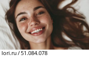 Smiling young Caucasian woman with long brown hair and freckles. She is lying on a bed, showcasing a joyful expression and natural beauty. Стоковое AI-видео, аниматор Nart / Фотобанк Лори