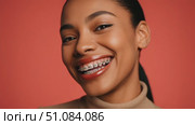 Smiling young African woman with braces, wearing a beige turtleneck against a vibrant orange background. She has dark hair styled in a bun and a joyful expression. Стоковое AI-видео, аниматор Nart / Фотобанк Лори