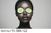A young African woman with short hair has cucumber slices over her eyes. She has a smooth, dark complexion and a serene expression. The background is neutral and minimalistic. Стоковое AI-видео, аниматор Nart / Фотобанк Лори