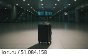 A black suitcase stands alone in an empty airport terminal. The floor is shiny and reflects the overhead lights. Digital flight information screens are visible in the background. Стоковое AI-видео, аниматор Nart / Фотобанк Лори