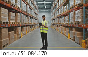 A young South Asian man stands in a warehouse aisle, wearing a yellow safety vest. He holds a tablet, surrounded by stacked cardboard boxes on shelves. Стоковое AI-видео, аниматор Nart / Фотобанк Лори