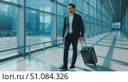 A young Caucasian man in a suit walks through a modern airport terminal, pulling a suitcase. Bright sunlight streams through large glass windows. Стоковое AI-видео, аниматор Nart / Фотобанк Лори