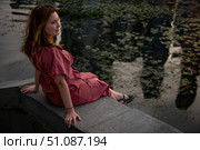 Woman in a burgundy dress sits on a stone embankment, gazing thoughtfully at the river surface with evening reflections. Стоковое фото, фотограф Софья Соломонова / Фотобанк Лори