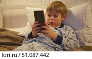 Cute kid boy in pajamas lying in bed watching smartphone, smiling. Children's screen time. Child plays games using phone at the evening or night. Social media addiction. Childhood, modern technology. Стоковое видео, видеограф Марина Демидюк / Фотобанк Лори