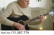 Senior man is playing guitar. Elderly man sitting on the sofa and playing guitar. Portrait of a gray-haired mature man in a sweater and glasses learning to play. Enjoying retirement life at home. Стоковое видео, видеограф Марина Демидюк / Фотобанк Лори