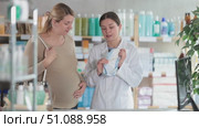 Friendly smiling female pharmacist in white coat explaining features of disposable baby diapers to interested pregnant woman, helping choose safe and comfortable option for future baby in pharmacy. Стоковое видео, видеограф Яков Филимонов / Фотобанк Лори