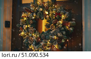 A beautifully decorated Christmas wreath hangs on a wooden door. The wreath features evergreen branches, pine cones, and warm lights, creating a festive winter atmosphere. Стоковое AI-видео, аниматор Nart / Фотобанк Лори