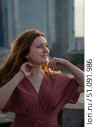 Smiling woman in a red dress touches her hair while standing outdoors against a blurred modern city background at sunset. Стоковое фото, фотограф Софья Соломонова / Фотобанк Лори