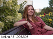 Woman in a rose dress sits on a bench near blooming hydrangeas, gazing thoughtfully into the distance with a serene expression. Стоковое фото, фотограф Софья Соломонова / Фотобанк Лори