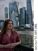 Woman in a red dress standing by a railing with Moscow City skyscrapers in the background at sunset. Редакционное фото, фотограф Софья Соломонова / Фотобанк Лори