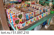 A bright counter with handmade souvenirs. Colourful photo frames decorated with seashells, pearls, decorative elements and inscriptions in Thai are neatly laid out on a wooden green table. Each frame is made in its own style: with hearts, stars, flowers and animals. In the background, plastic baskets with other goods are visible, as well as elements of the interior of the shop. The atmosphere of the photo conveys the colour of a traditional market and the variety of products of local craftsmen. Стоковое фото, фотограф Анна Кривицкая / Фотобанк Лори