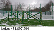 Outdoor sports ground in winter. In the center of the frame are green metal structures with a camouflage pattern, designed for training, overcoming an obstacle course or physical training. The structure consists of crossbars intersecting at an angle, fixed to vertical posts. The site is fenced with a high metal fence, behind which you can see bare trees and snowdrifts. On the ground is an artificial green surface, partially covered with snow. The atmosphere of the photo combines winter and a sporty spirit, demonstrating the conditions for outdoor activities. Стоковое фото, фотограф Анна Кривицкая / Фотобанк Лори