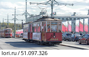A photo of an old retro tram on the streets of St. Petersburg. A red tram with wooden window frames and the number 2424 is traveling across a bridge, with other trams of the same era visible behind it. In the background, you can see the city's architecture with classical facades, as well as festive flags with the St. George ribbon fluttering in the wind. There are many cars and passers-by on the street, creating the atmosphere of the city. The photo conveys the spirit of history and nostalgia, recalling the old transport that was once the main means of transportation in the city. (2015 год). Редакционное фото, фотограф Анна Кривицкая / Фотобанк Лори