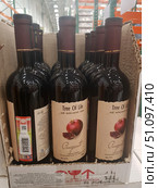 Cardboard box with bottles of Tree Of Life pomegranate wine. Red semi-sweet wine, 12% strength, produced in Armenia. The label depicts a juicy pomegranate with grains, the brand name and product information in English. The bottles have dark glass and are decorated with burgundy corks with a golden stripe. The photo was taken in a store or warehouse - metal shelves and boxes are visible in the background. The atmosphere of the photo emphasizes the naturalness and origin of the drink, associated with the traditions of Armenian winemaking. (2019 год). Редакционное фото, фотограф Анна Кривицкая / Фотобанк Лори