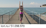 Woman jogging towards camera and performing dynamic warm-up arm movements on concrete pier by the sea under bright morning sunlight, front view fitness routine promoting energy and healthy lifestyle. Стоковое видео, видеограф Сергей Тимофеев / Фотобанк Лори