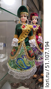 Souvenir dolls in national costumes. In the foreground is a doll in a green dress with a bright pattern and gold decorations. The skirt is decorated with floral and oriental patterns in green and blue tones, complemented by lace and gold decor. On the chest is a large decorative red stone, framed with gold embroidery. The headdress is made of green velvet and decorated with gilded elements and pendants. In the background is another doll in a burgundy outfit with similar elements, but with a purple stone. Such dolls reflect ethnic traditions, the richness of national costumes and serve as popular souvenirs. Стоковое фото, фотограф Анна Кривицкая / Фотобанк Лори