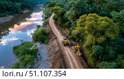 Aerial view of jungle road construction alongside serene river at sunset. Стоковое AI-изображение, AI-художник Olga Didraga / Фотобанк Лори