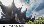 Ancient Chinese architecture. A fragment of a building. Tiled roofs with curved eaves against a blue sky, clouds. Latticed wooden balcony railings. Green vegetation. China. Shanghai. Yu Garden. Yuyuan. Стоковое фото, фотограф Вера Ивановна Кубарева / Фотобанк Лори