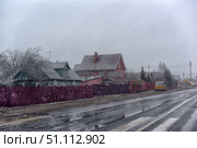 A winter rural landscape with houses along the road. In the foreground is an asphalt road with markings, wet from melting snow, with puddles and reflections. In the background are private houses: a green wooden house with antennas and satellite dishes and a large brick house with a roof covered with snow. Behind the houses there is a fence of a rich burgundy color. Snow is falling, noticeable snowflakes create an atmosphere of winter bad weather and coziness at the same time. On the right is parked a yellow bus, along the street there are road signs and a traffic light. The sky is covered with gray clouds, which emphasizes the cold and cloudy winter day. The scene conveys the atmosphere of the Russian outback and real life in a small village or town. Стоковое фото, фотограф Анна Кривицкая / Фотобанк Лори