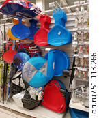 Shelves in a store with winter sports goods. In the foreground are various sleds and snow scooters for children: bright plastic round and oval sleds, red and blue scoop sleds with a handle, round tubes with cartoon characters, as well as plate sleds with a steering wheel. All the goods are neatly hung and laid out, creating a bright and color-saturated composition. Skis are visible on the left edge of the frame, and part of a display case with fishing goods is on the right. The photo conveys the atmosphere of winter shopping and preparation for active recreation in the fresh air. (2019 год). Редакционное фото, фотограф Анна Кривицкая / Фотобанк Лори