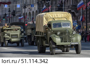 Military equipment from the Second World War on parade in the city. In the foreground is an army truck in protective green paint with a tarpaulin body. The vehicle is equipped with a characteristic grille on the radiator, and the number plate says "LENRESERVE". Other military vehicles are visible behind it, including a half-track armored personnel carrier. The column moves along a city street decorated with Russian flags, with spectators gathered on both sides. The scene recreates the atmosphere of military history and the memory of the Great Patriotic War. (2015 год). Редакционное фото, фотограф Анна Кривицкая / Фотобанк Лори