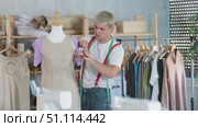 Tailor stands in the workshop near a mannequin with a measuring tape. Male fashion designer is working on a collection of clothes and stands against the background of shelves and racks with fabrics. Стоковое видео, видеограф Яков Филимонов / Фотобанк Лори