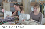 Two female tailors are sitting at a table in the workshop and sewing clothes on machines. Employees of the sewing industry make wear according to individual measurements. Стоковое видео, видеограф Яков Филимонов / Фотобанк Лори