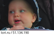 Close-up of the face of a cute full-cheeked baby sitting in a stroller. The baby has cute blue eyes and two bottom teeth. Happy baby boy wearing an adult baseball cap. Стоковое видео, видеограф Воловодов Владимир / Фотобанк Лори