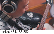 A person is immersed in playing guitar, wearing large headphones. They sport a stylish plaid shirt, surrounded by a cozy indoor setting, suggesting a creative atmosphere. Guitar work in a home studio. Стоковое видео, видеограф Воловодов Владимир / Фотобанк Лори