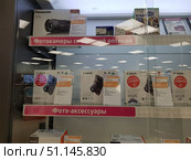 A display case of an electronics store with cameras and video cameras. The top shelf contains boxes with equipment, including a Panasonic HC-V800, as well as Instax film and Transcend memory cards. The middle shelf contains boxes with Canon IXUS and PowerShot cameras with price tags and promotional stickers "As a gift!" The price tags with the characteristics and cost of the models are visible. The bottom shelf contains accessories and compact Sony cameras. The display case is decorated with dividers with the inscriptions "Cameras with interchangeable optics" and "Photo accessories". The photo conveys the atmosphere of a modern electronics store, a wide selection of photo products and accessories. (2019 год). Редакционное фото, фотограф Анна Кривицкая / Фотобанк Лори